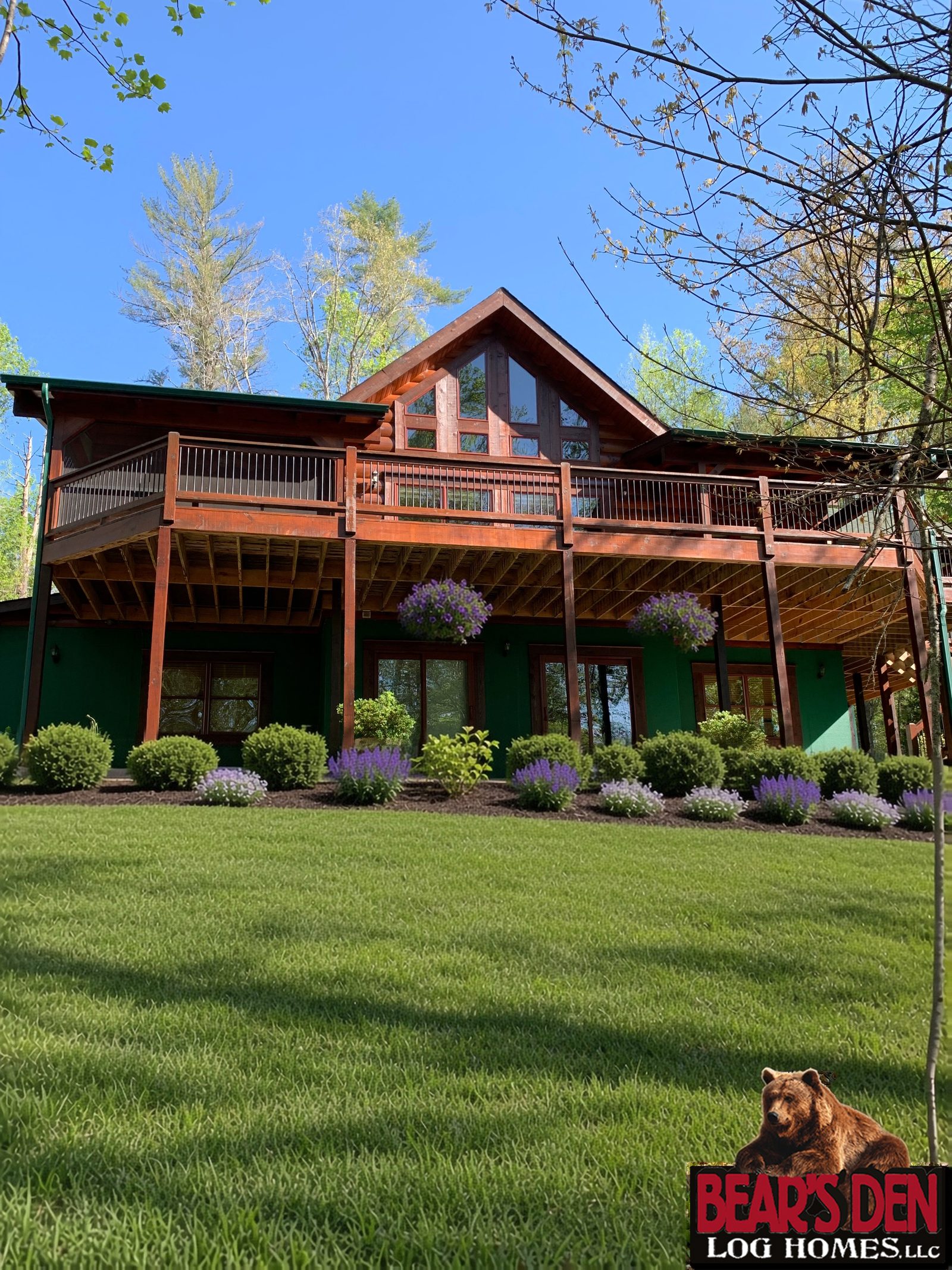 Log cabin with a wraparound deck and tall gable windows above a landscaped bed and a sloping green lawn