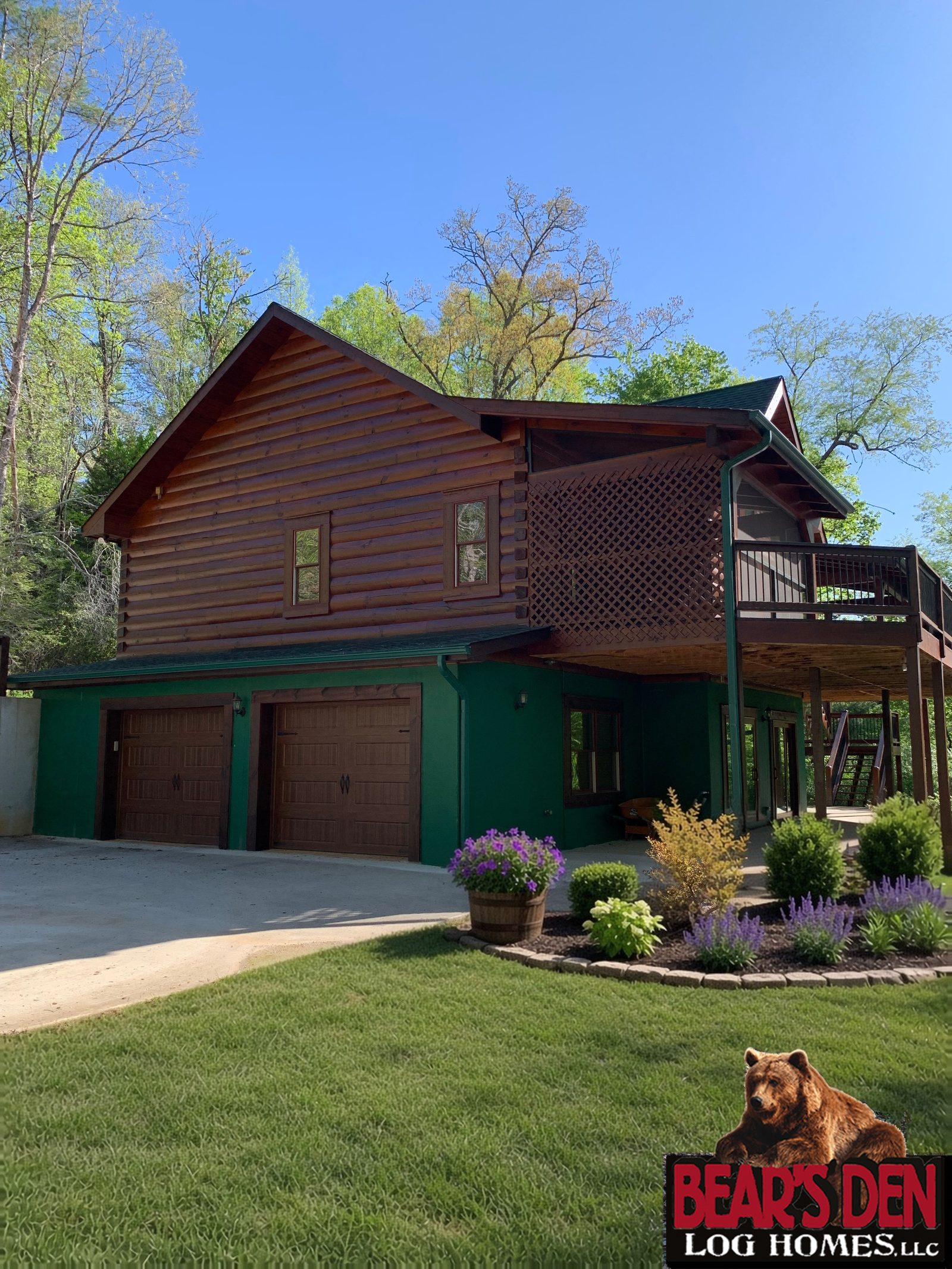 Log cabin with a lower level garage, a stone edged flower bed with purple salvia and a barrel planter of petunias