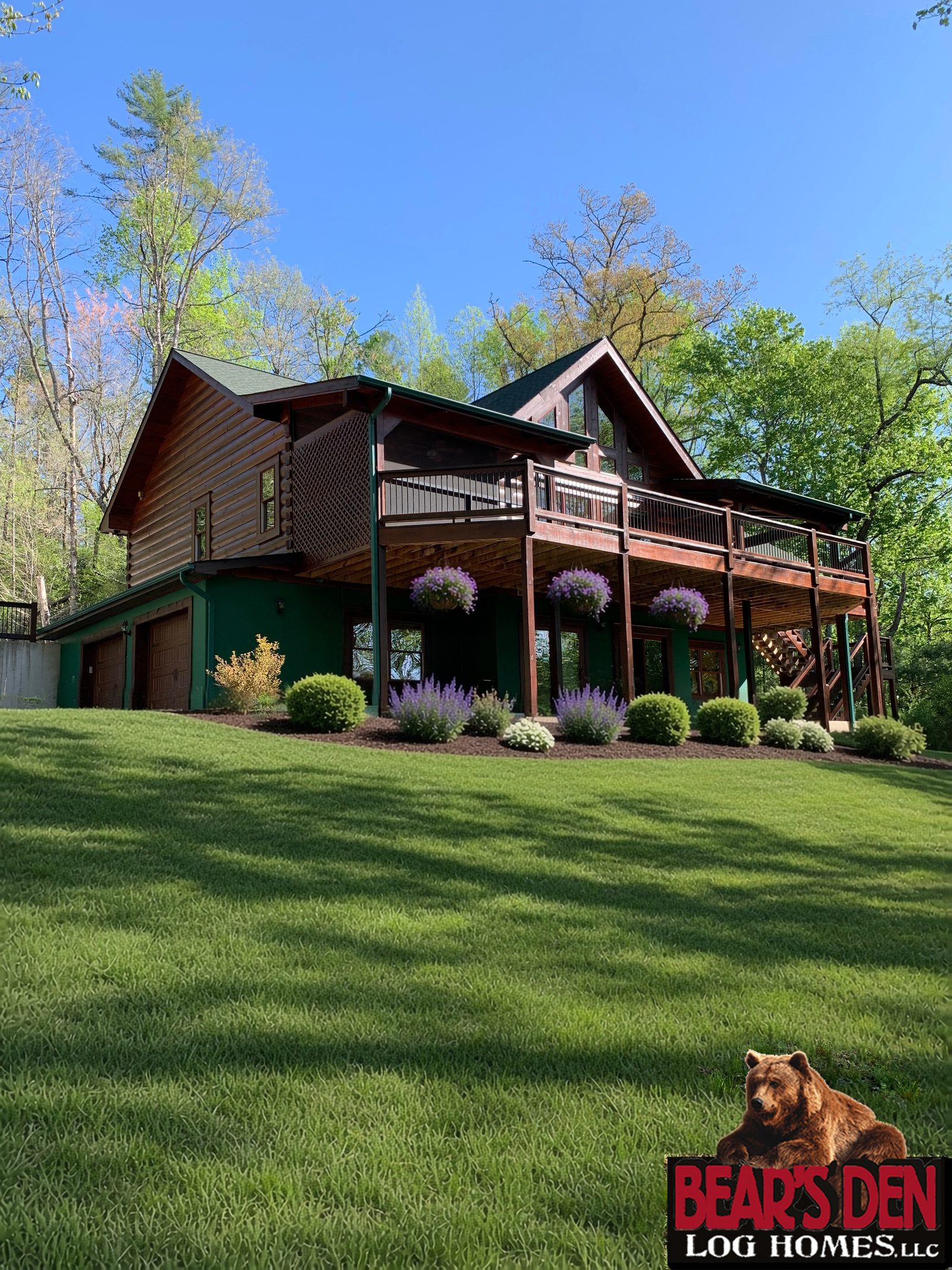 Log cabin on a wooded hillside with a covered deck, hanging flower baskets and landscaped beds of boxwood and purple salvia above a green lawn